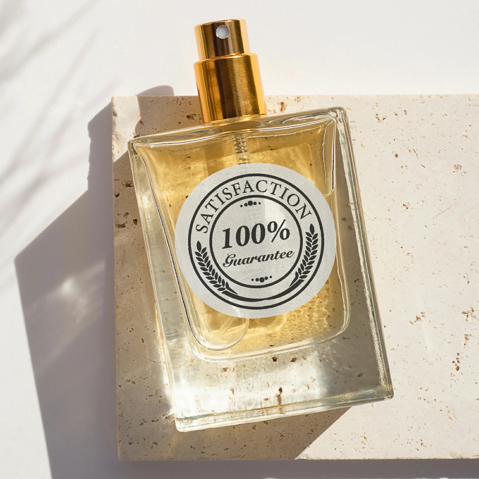 Transparent perfume bottle with gold spray top and round '100% Satisfaction Guarantee' thermal label on stone surface