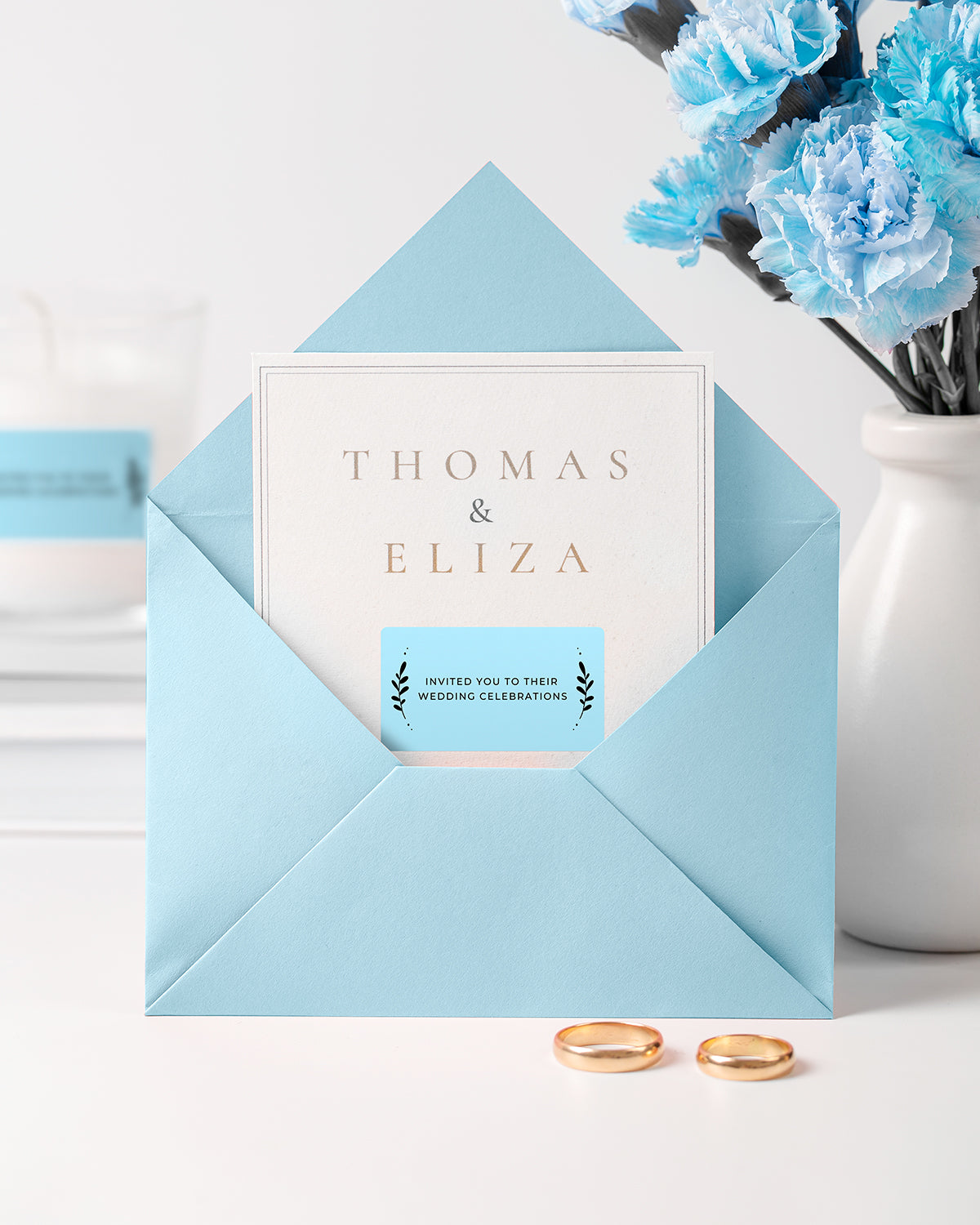 Blue envelope with wedding invitation, gold rings, blue flowers, and candle on white background