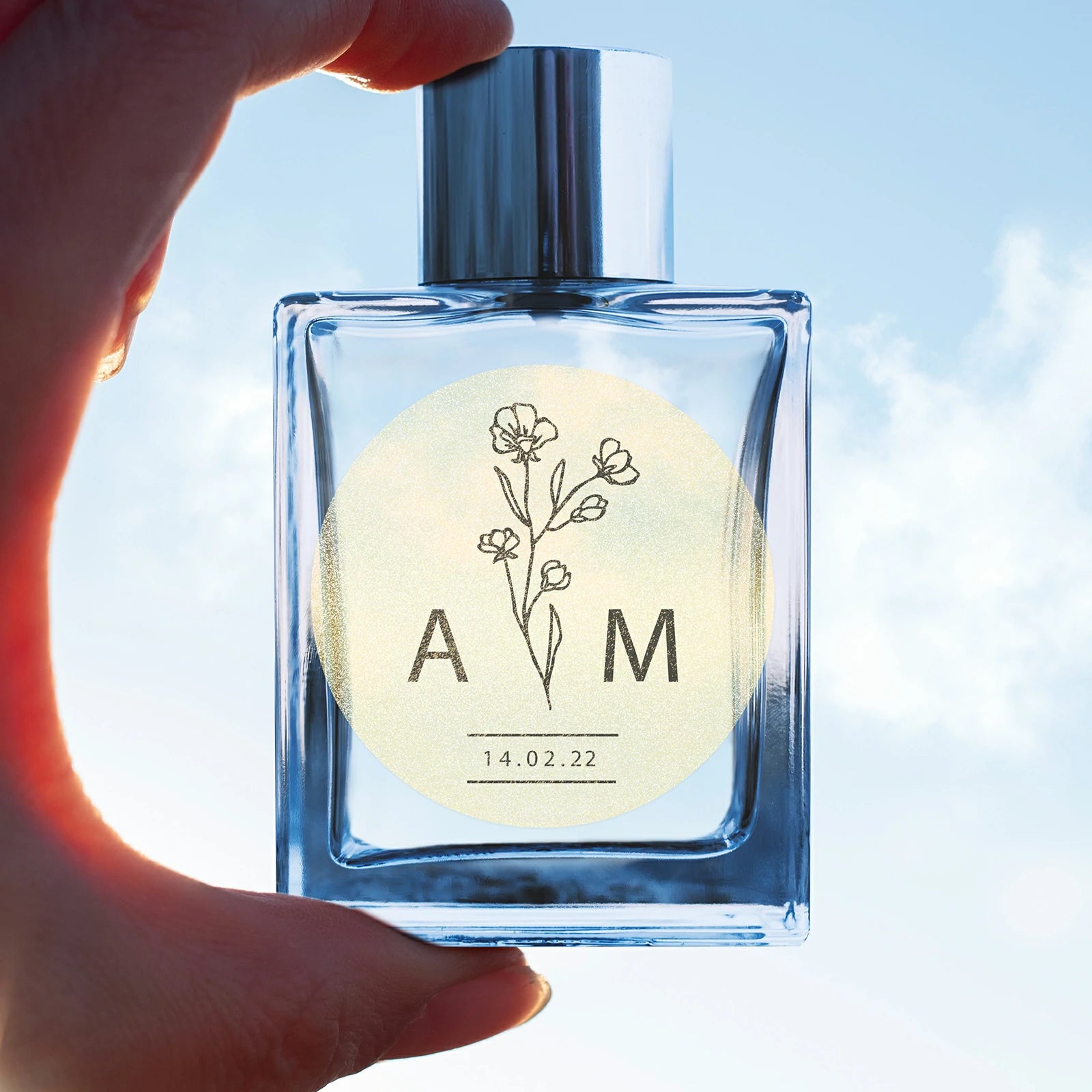 Hand holding clear glass bottle with floral thermal label, personalized monogram and date.