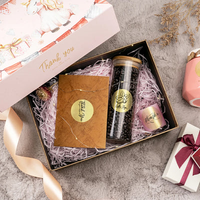 Gift box with thank you label, glass coffee jar, card, and pink candle on shredded paper.