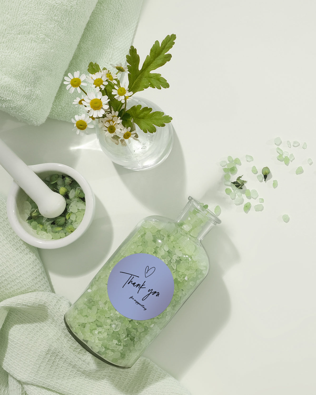 Glass jar of green bath salts with thank you label, chamomile flowers, and textiles on white background