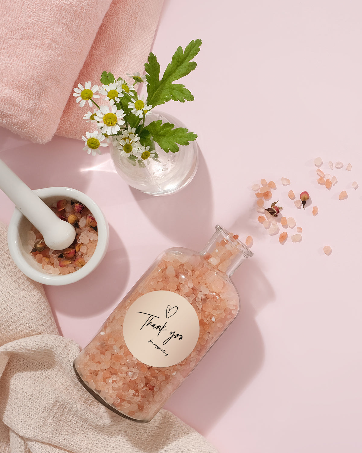 Spa flat lay with pink bath salts in glass jar, chamomile flowers, mortar and towel on pink background