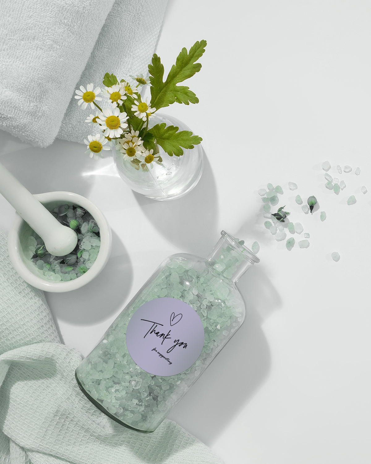 Glass jar of green bath salts with thank you label, chamomile flowers, and spa towels
